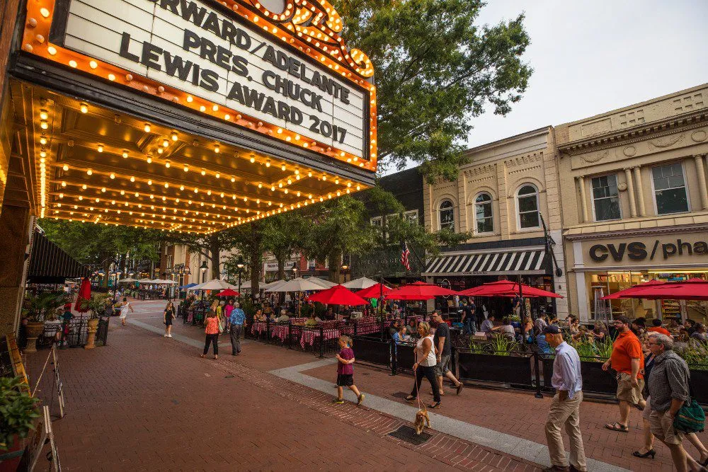 A bustling street scene with outdoor diners under red umbrellas, bright marquee lights, and shops lining the vibrant sidewalk.