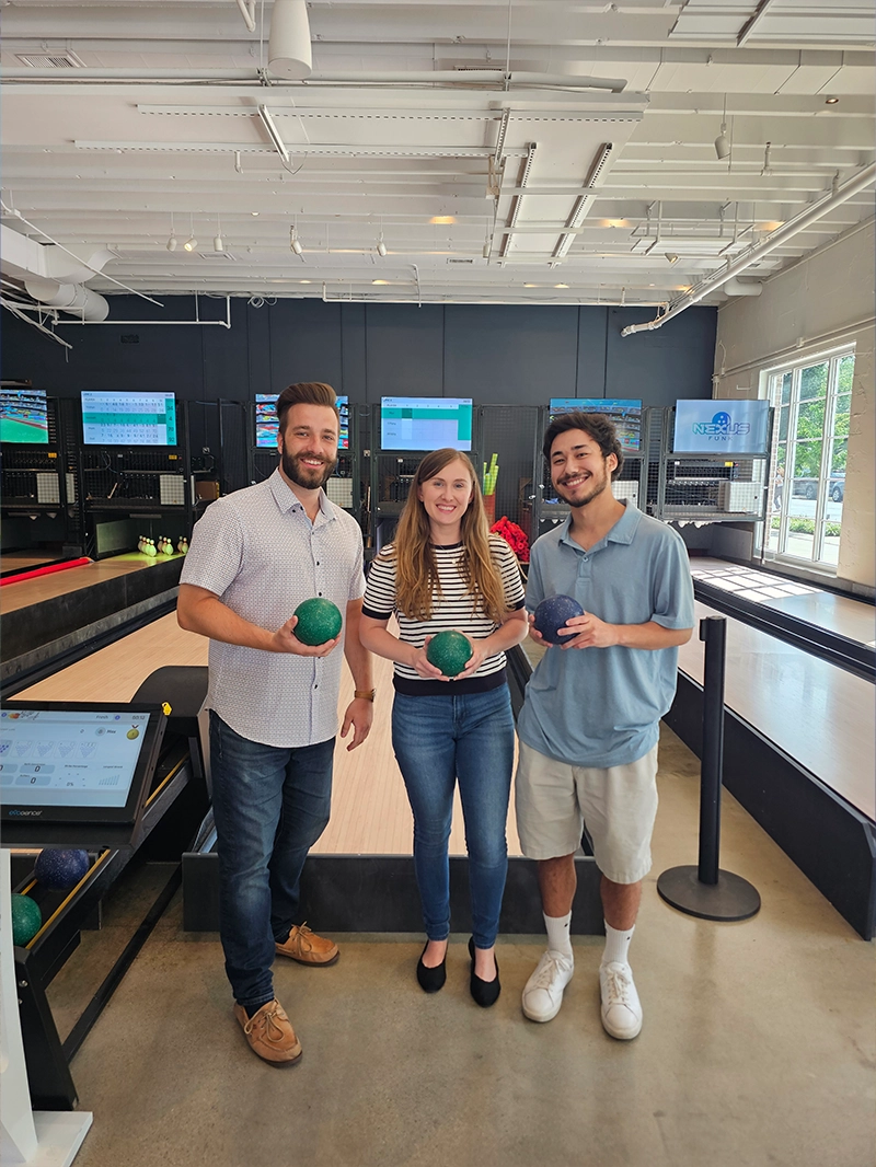 Three individuals stand together holding bowling balls in a modern bowling facility, with screens displaying game information in the background.