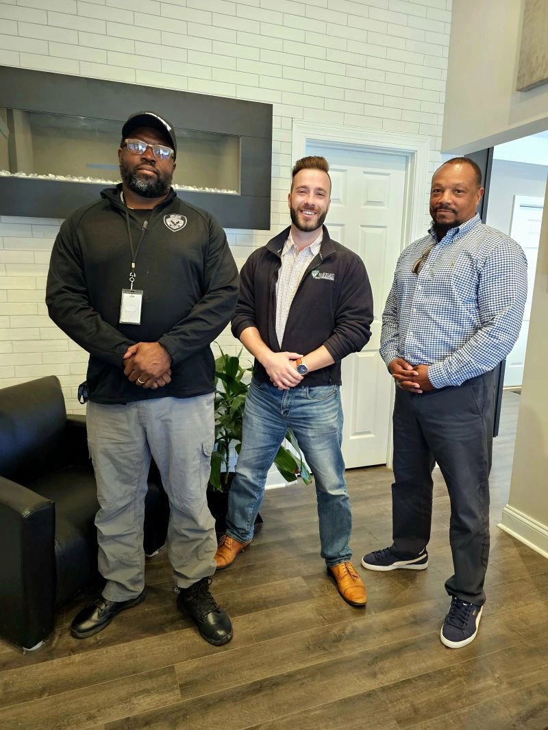Three men standing in an office, positioned in front of a couch, engaged in conversation